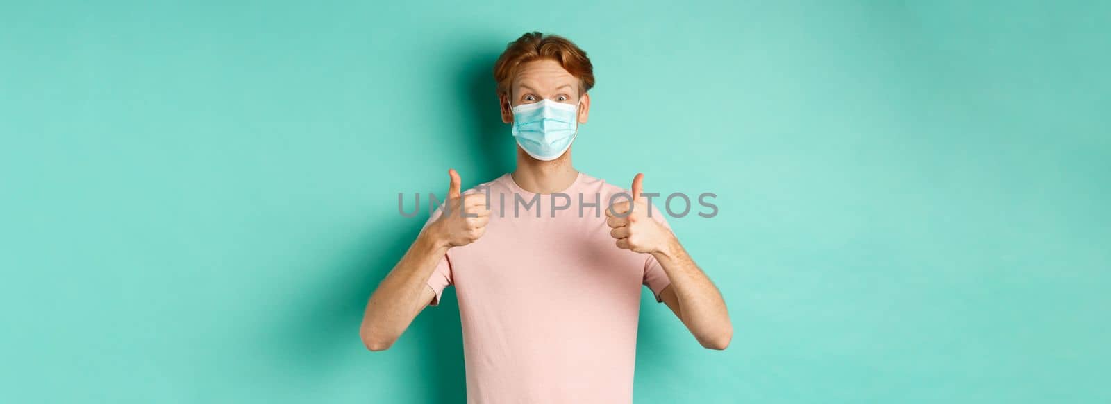 Covid-19, pandemic and lifestyle concept. Cheerful redhead guy in medical mask showing thumbs up in approval, like and praise product, standing over turquoise background.