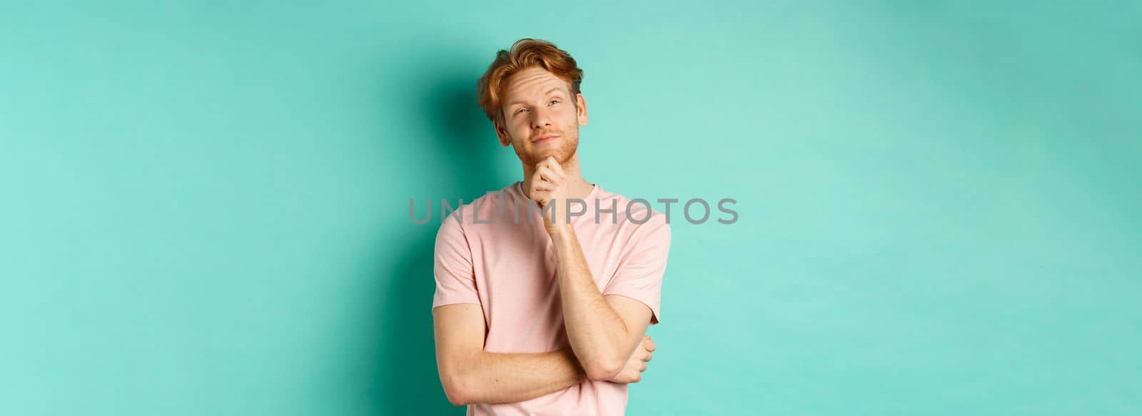 Pensive handsome man with red hair and beard looking at upper left corner, making choice and looking thoughtful, standing in t-shirt over mint background.