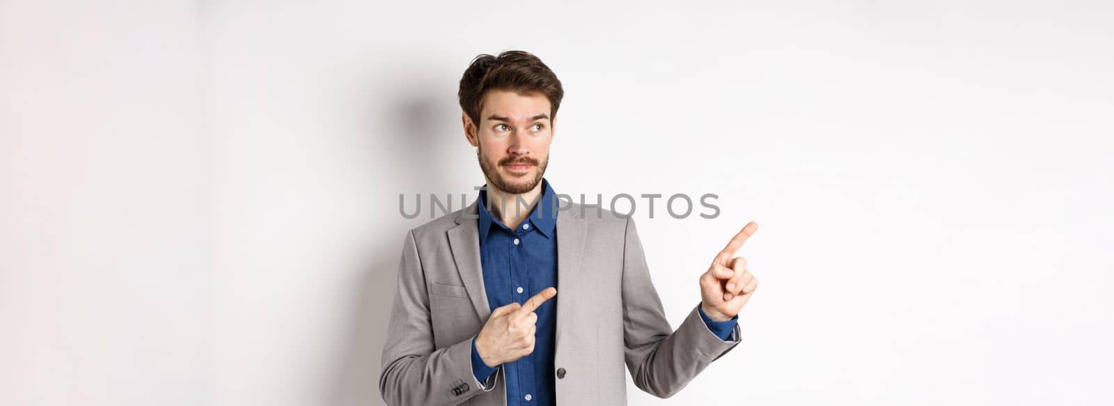 Doubtful male entrepreneur in suit looking and pointing left with hesitant face, standing unsure against white background.