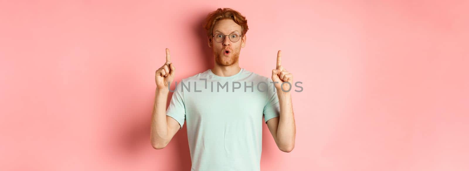 Impressed caucasian man with ginger hair, wearing glasses and t-shirt, saying wow and pointing fingers up at awesome deal, standing over pink background.