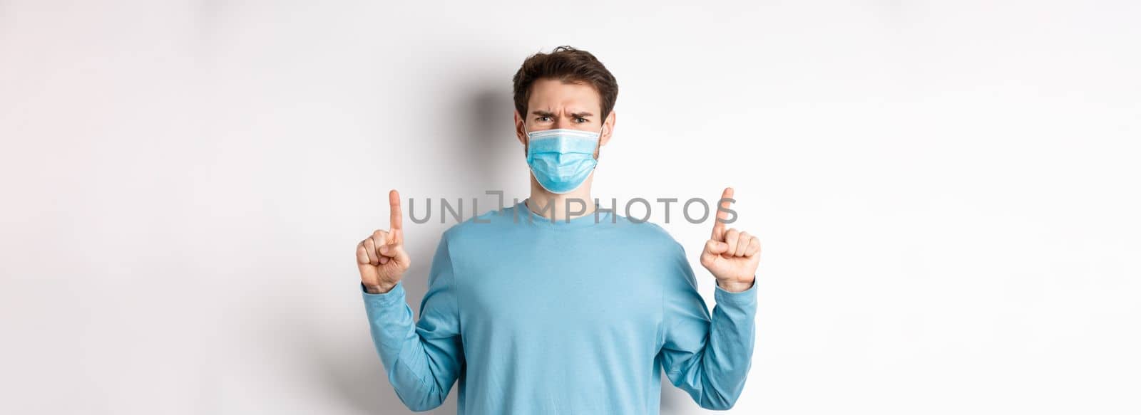 Coronavirus, health and quarantine concept. Disappointed young man frowning at absurd, pointing fingers up and complaining at bad promo, wearing medical mask, white background.