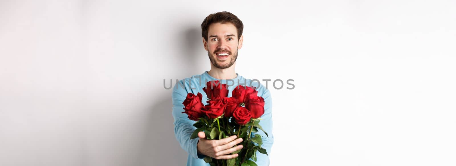 Handsome bearded guy stretch out hands, giving bouquet of roses and smiling, bring flowers on romantic date, celebrating Valentines day with lover, standing over white background.