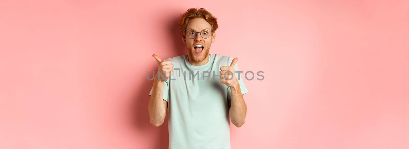 Amazed redhead guy looking impressed and excited, showing thumbs up in approval, praise awesome promo offer, standing over pink background by Benzoix