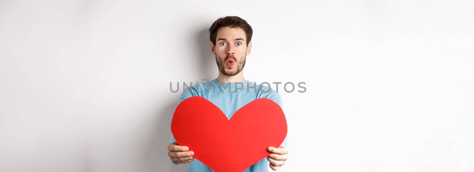 Young handsome man showing big red valentines day heart, being in love, pucker lips for kiss on valentine date, standing over white background.