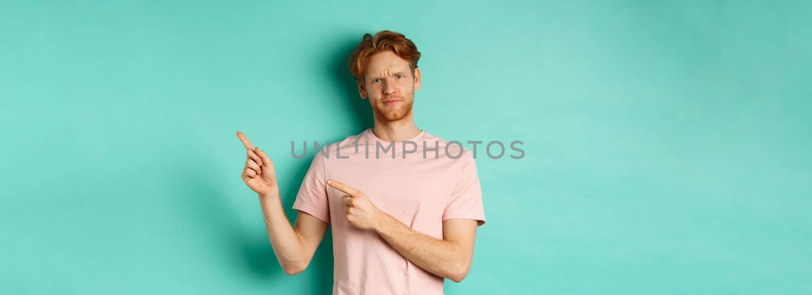 Skeptical and doubtful redhead man in t-shirt pointing fingers at upper right corner, showing promo offer with displeased face, standing over turquoise background.