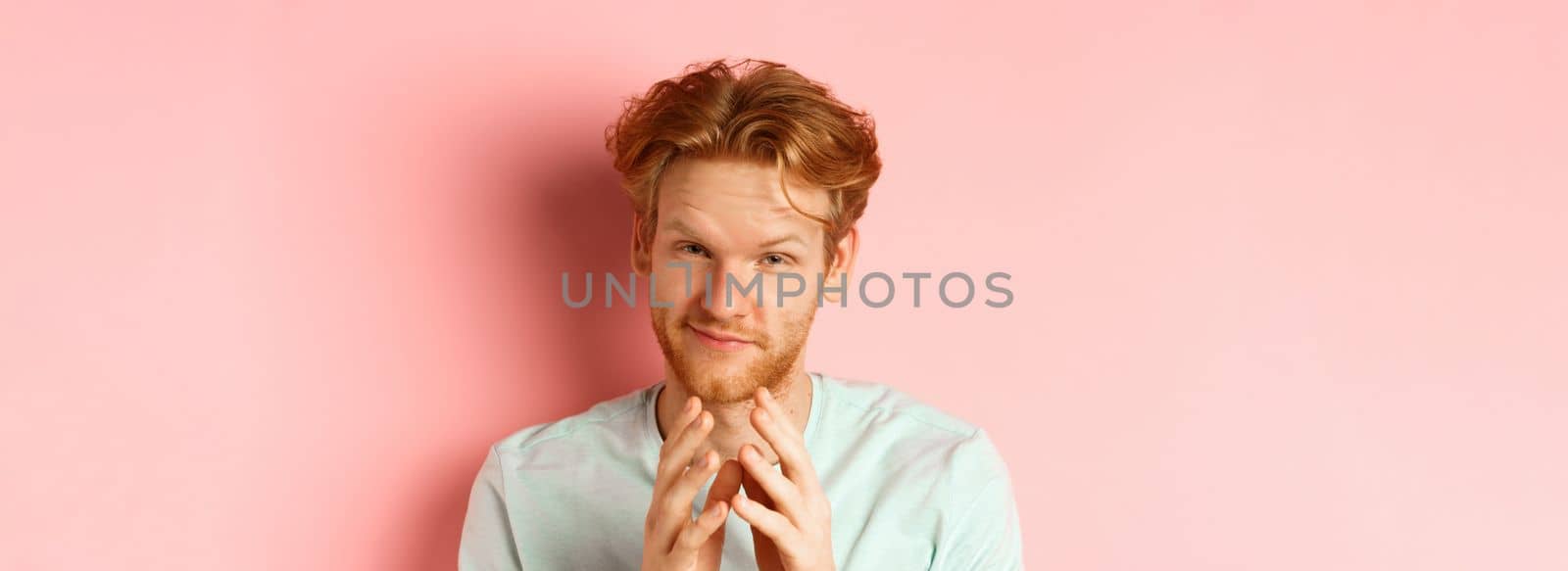 Close up of funny bearded man with red hair pitching a perfect plan, smiling and steeple fingers, scheming something, standing devious against pink background.