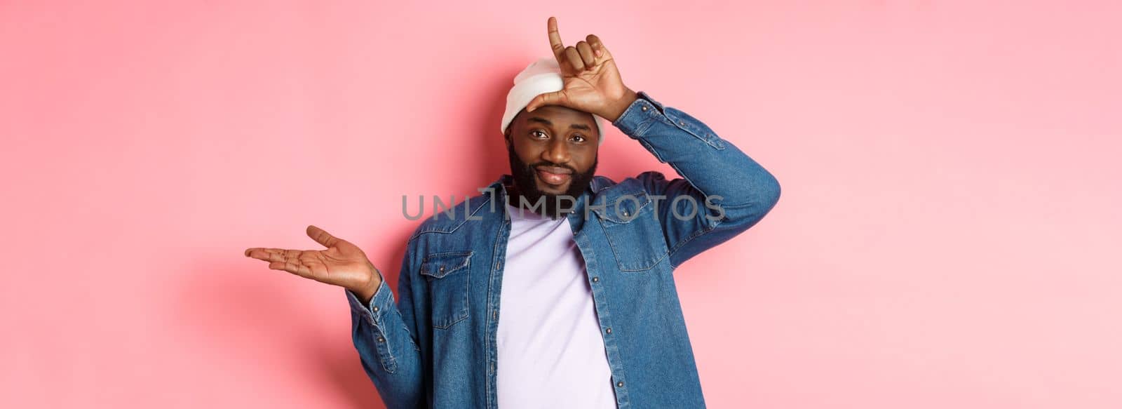 Sad african-american man showing loser sign on forehead and staring at camera, standing over pink background.