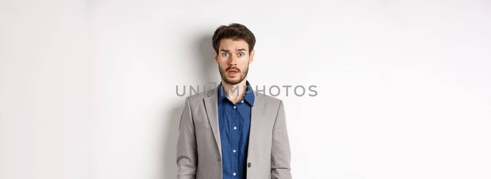Shocked male entrepreneur in suit drop jaw, staring worried and confused at camera, standing against white background.