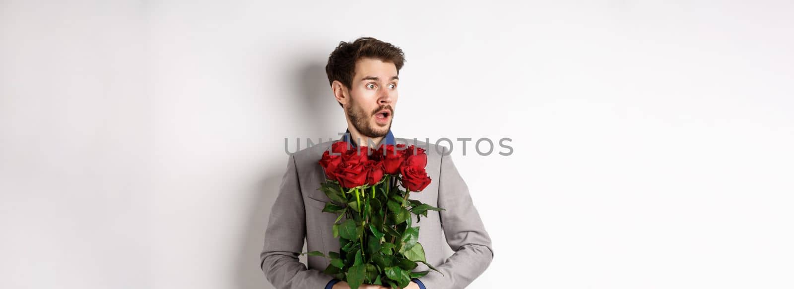 Handsome young man in suit holding red roses, looking left with surprised and startled expression, standing on Valentines day over white background.
