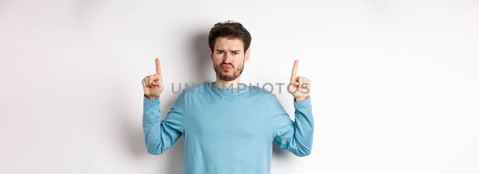 Disappointed male model frowning and sulking, pointing fingers up with negative emotion, dislike promo offer, standing over white background.