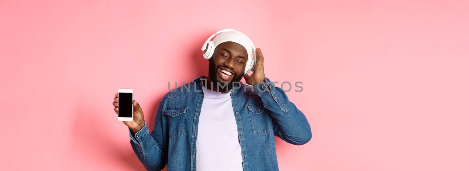 Carefree handsome Black man listening music in headphones, enjoying sound and showing smartphone screen, standing with pleased smile on face against pink background.