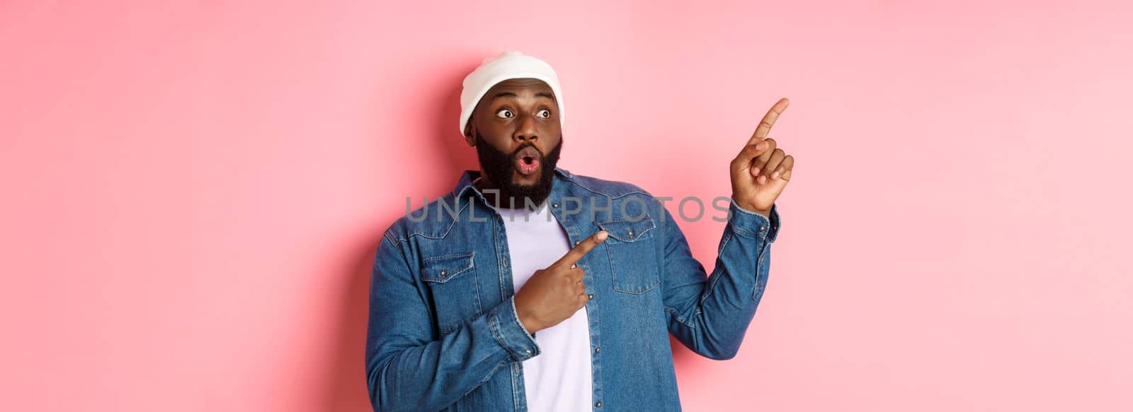 Impressed hipster black man in beanie and denim shirt, looking and pointing at upper left corner logo, staring amazed, standing over pink background.