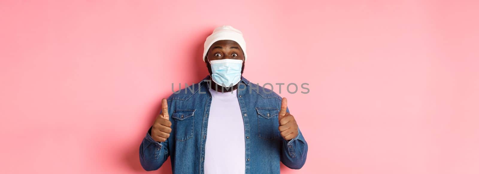 Covid-19, lifestyle and quarantine concept. Satisfied Black man in face mask showing thumbs-up, standing over pink background.