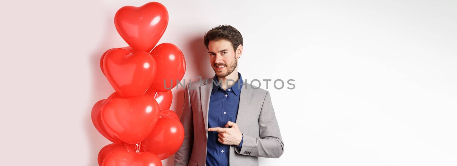 Lovers day. Charming young man with beard, wearing fancy suit, pointing finger at heart balloon surprise for Valentines day, standing over white background by Benzoix