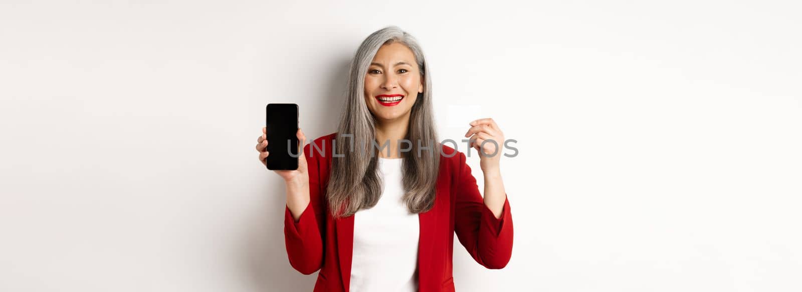 Senior asian businesswoman showing plastic credit card and blank smartphone screen, smiling at camera, white background.