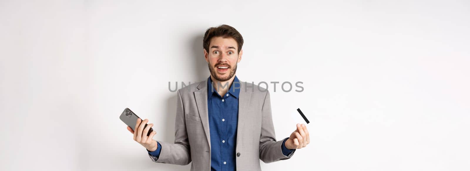 Online shopping. Surprised businessman holding plastic credit card and smartphone, making money in internet, standing in suit against white background by Benzoix