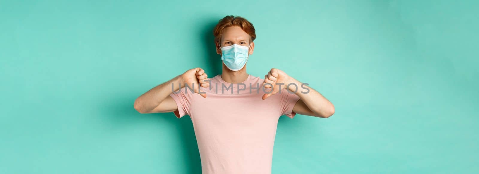 Covid-19, pandemic and lifestyle concept. Young man with red hair in face mask, showing thumbs down, dislike or disapprove something, standing over turquoise background.