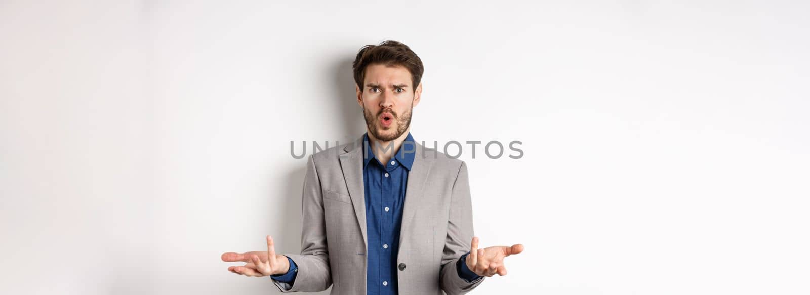 Confused and shocked businessman raising hands up and asking what happened, frowning disappointed, waiting for explanation, standing in suit on white background.