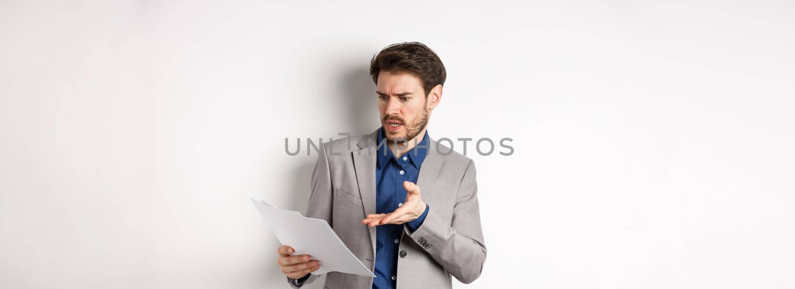 Angry employer looking at bad document, complaining on deal, pointing at paper frustrated, standing in suit on white background.