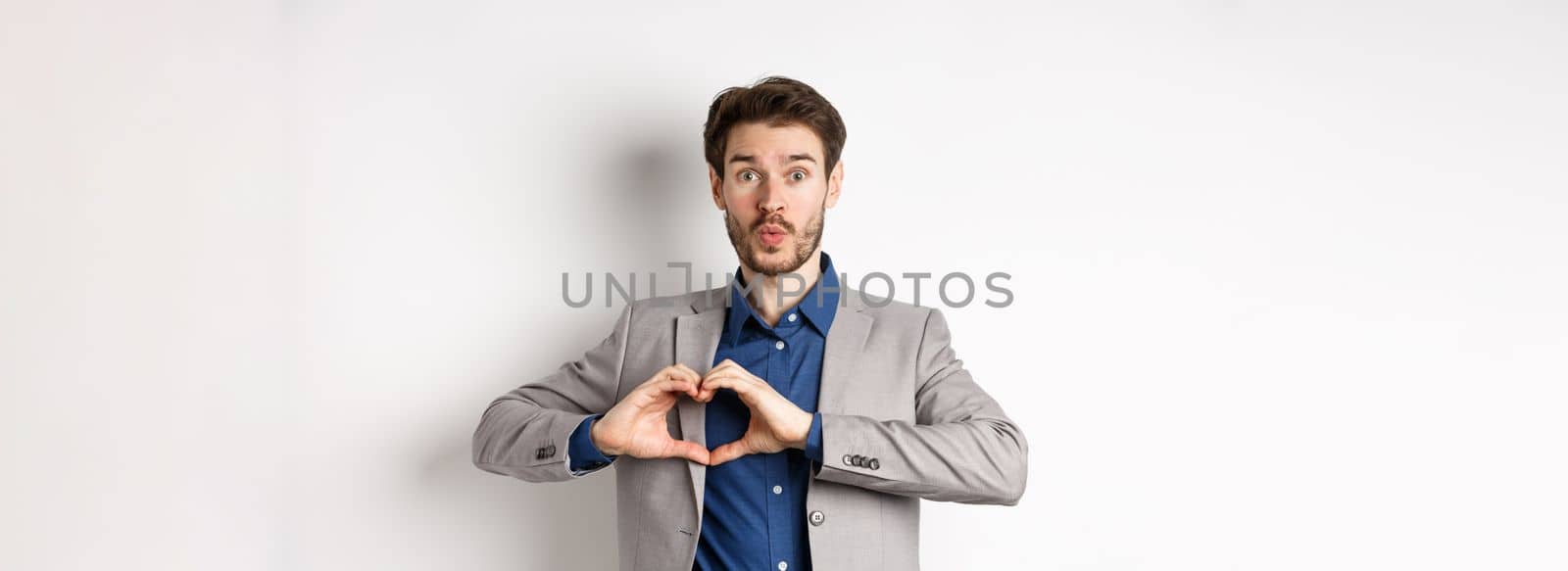 Handsome man looking silly, showing heart love you sign and pucker lips, waiting for kiss from lover, standing in suit on white background.
