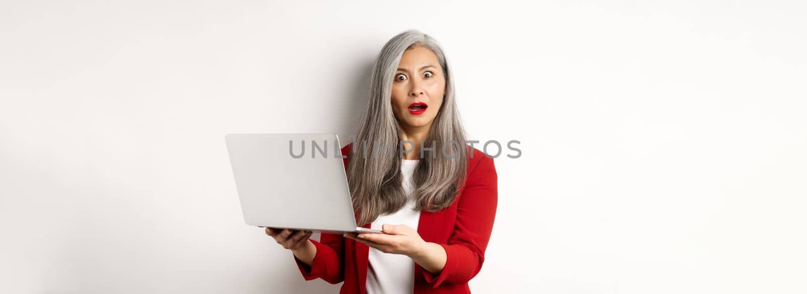 Business. Impressed asian old woman working on laptop, staring at camera surprised, standing over white background by Benzoix