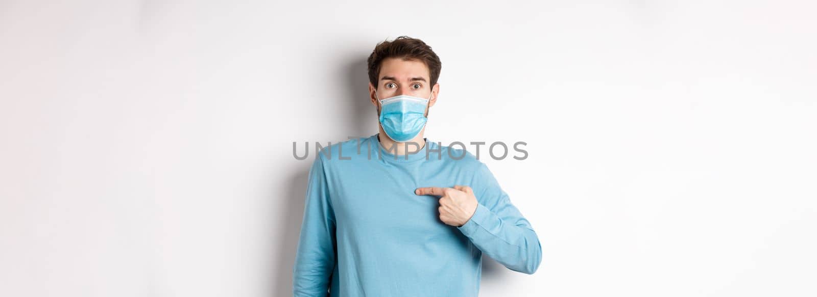 Coronavirus, health and quarantine concept. Surprised guy in medical mask points at himself, standing over white background.