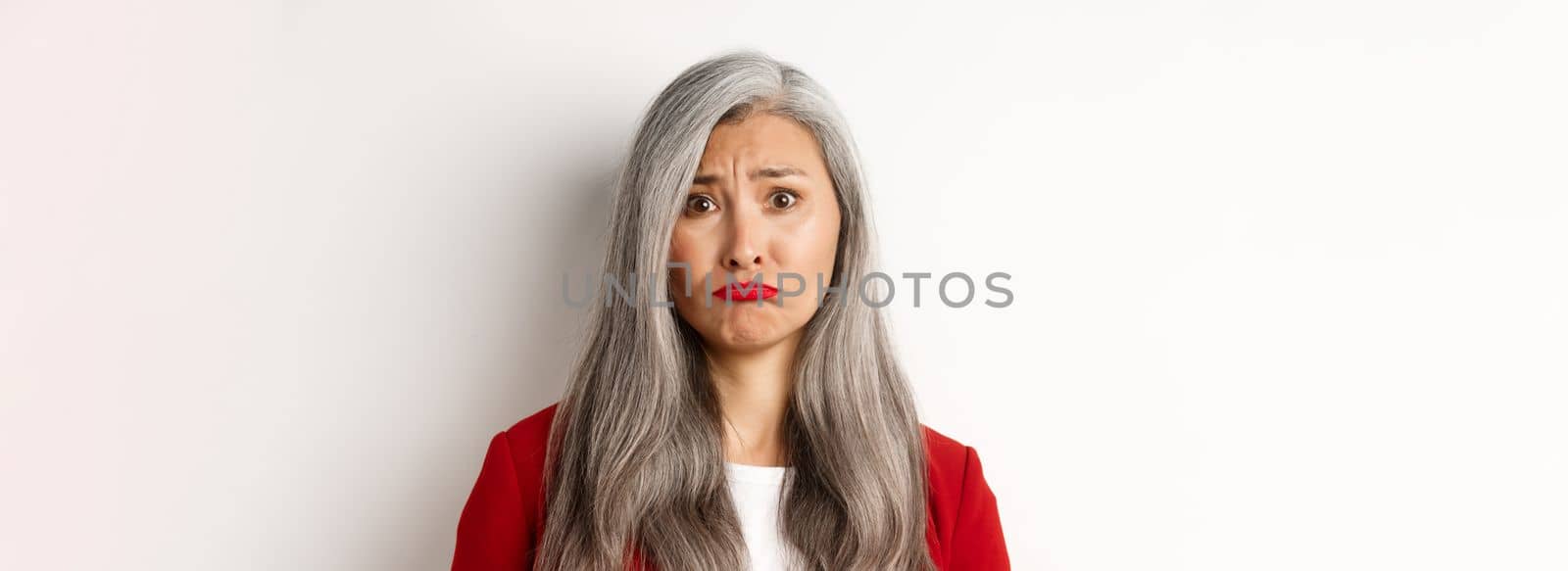 Close up of sad and unhappy asian senior woman sulking, frowning and looking jealous at camera, standing over white background.