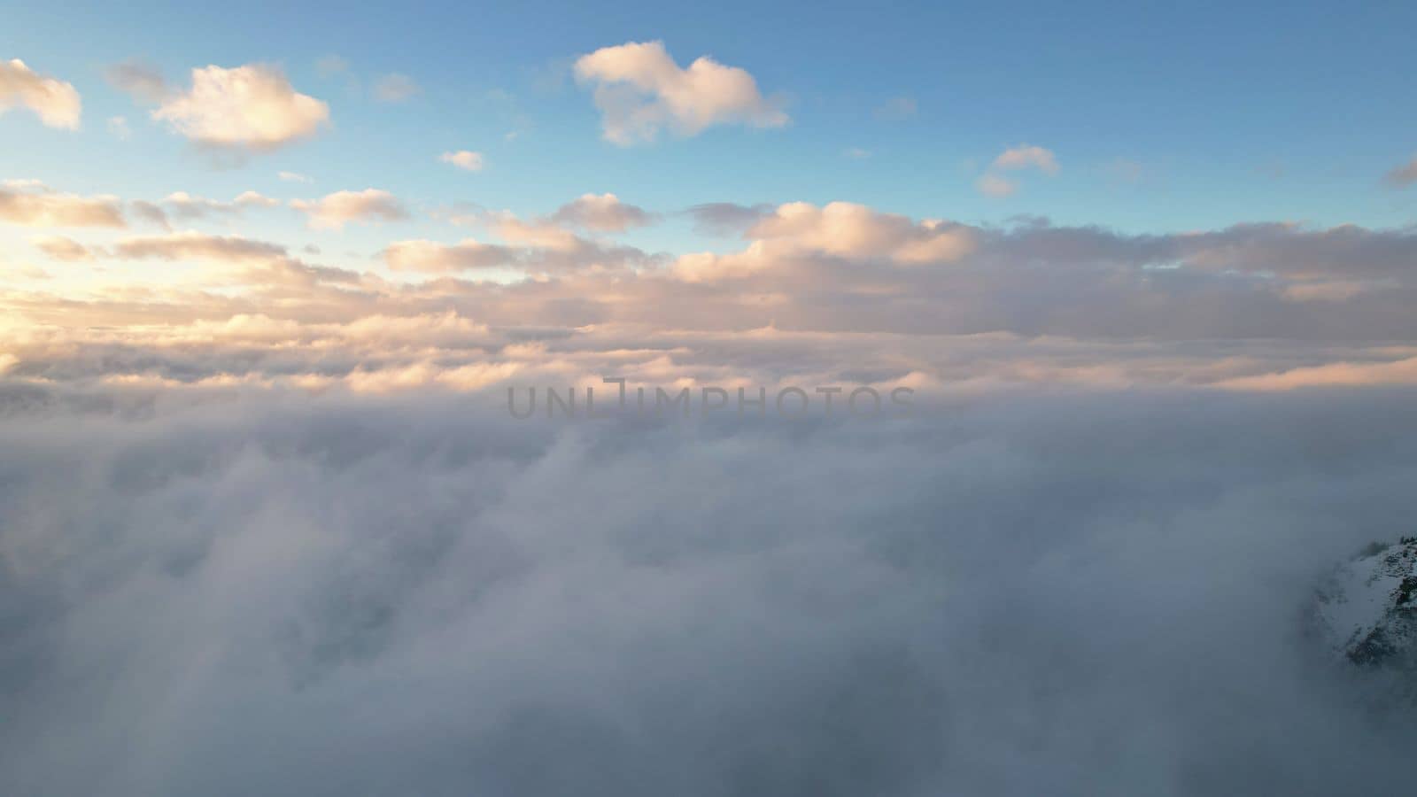 Flying among the ocean of clouds in the mountains. The fog is like big ocean waves crashing against high mountains covered with snow. Spruce trees grow in places. Sunset. Orange rays of the sun