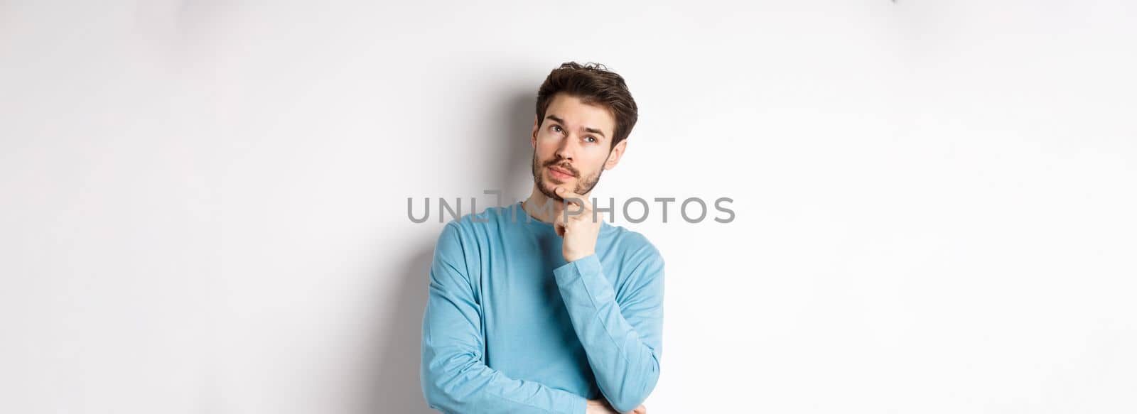 Image of handsome young man making choice, thinking and looking pensive up, standing over white background.