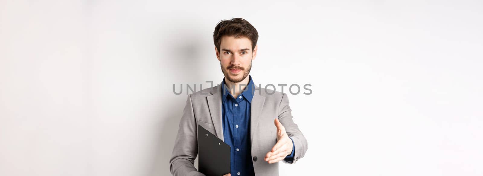 Confident businessman with clipboard giving hand for handshake, greeting business partners on meeting, standing on white background in suit.