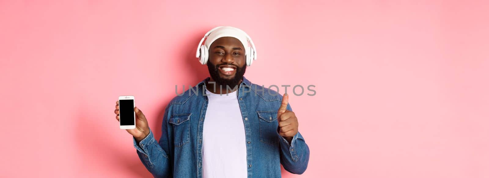 Happy hipster guy listening music on headphones and showing mobile screen, smiling satisfied, showing thumb-up in approval, like playlist, standing over pink background.