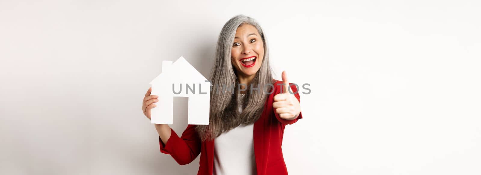 Real estate. Portrait of asian female broker showing thumb-up and paper house cutout, recommending agency to buy property, standing happy over white background by Benzoix