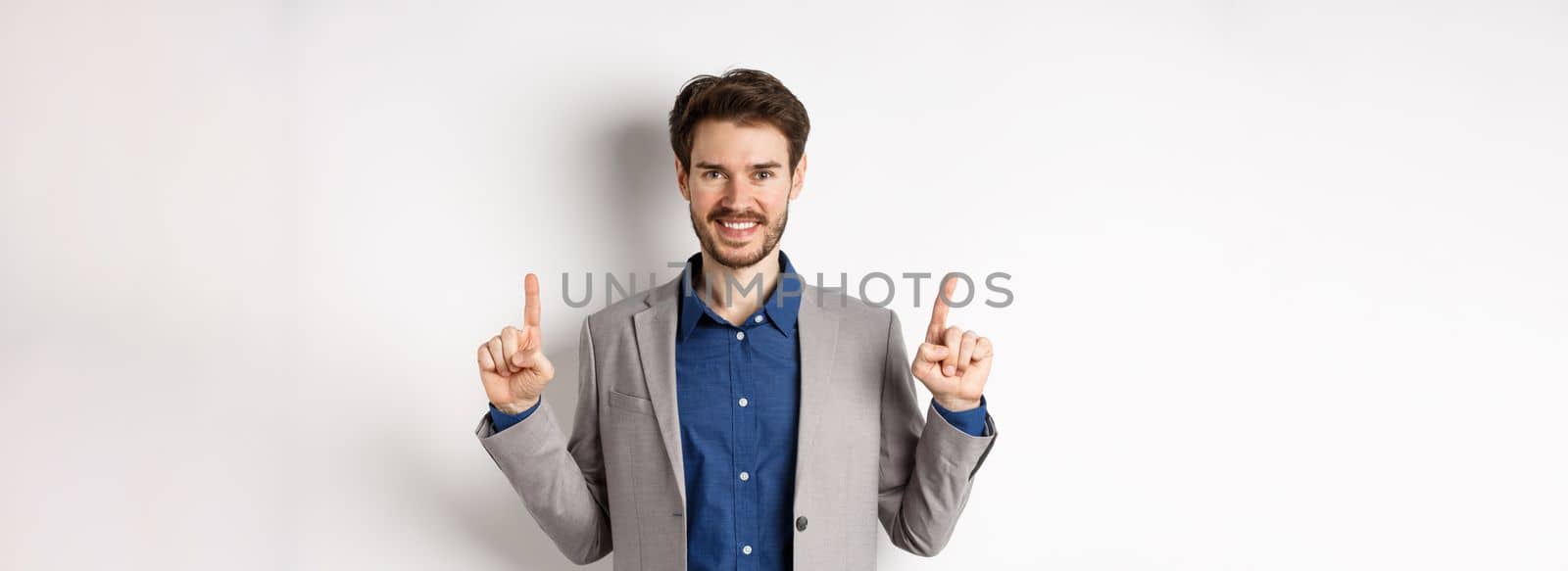 Confident successful businessman pointing fingers up, smiling and showing company logo, standing in suit on white background.