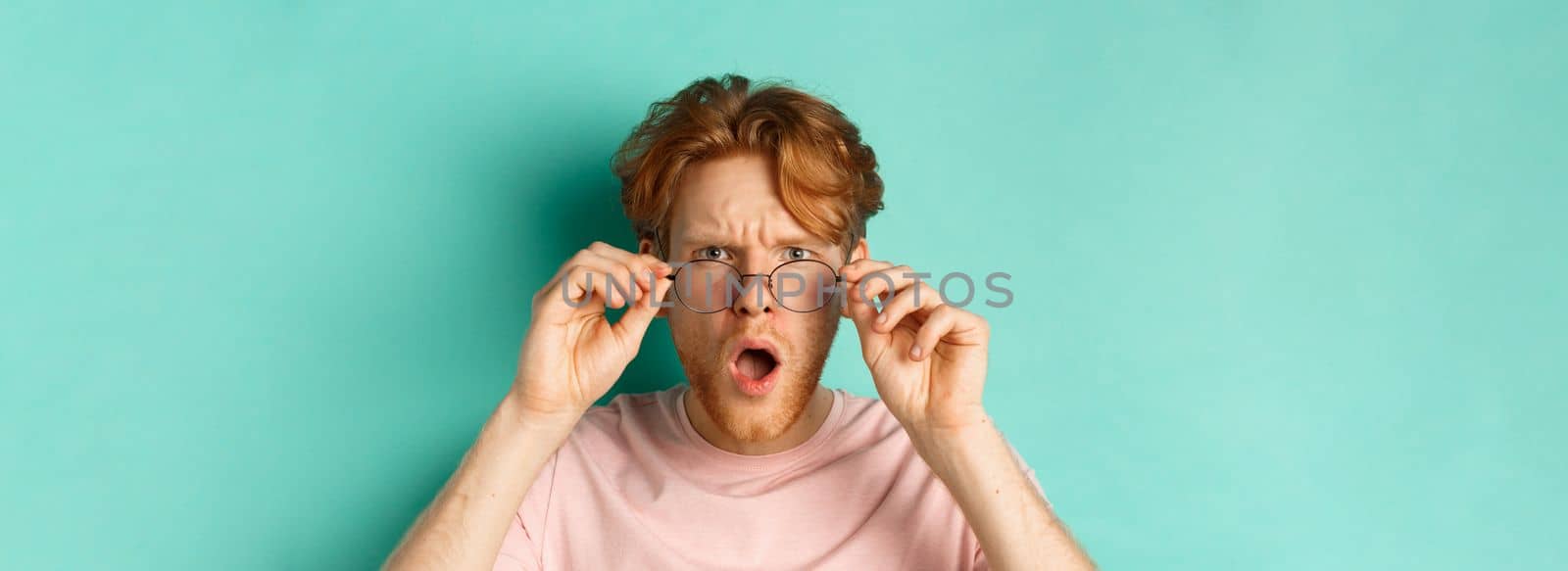Close up portrait of redhead guy take-off glasses and gasping startled, looking shocked at camera, standing over turquoise background.