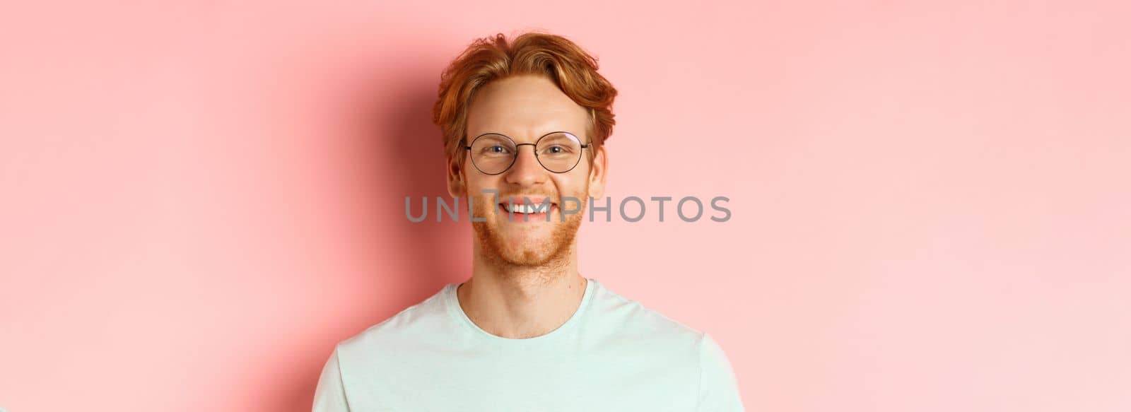 Close up of happy redhead man face, smiling with white teeth at camera, wearing glasses for better sight and t-shirt, standing over pink background.