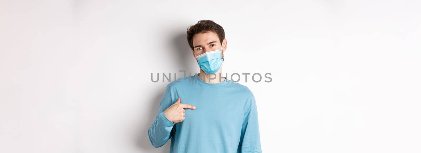 Covid-19, health and quarantine concept. Smiling caucasian man in medical mask pointing at himself, volunteering, standing over white background.