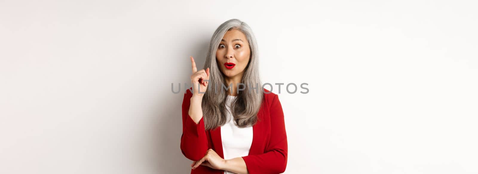 Elderly asian woman entrepreneur in red blazer having an idea, suggesting something, raising finger in eureka gesture, standing over white background by Benzoix