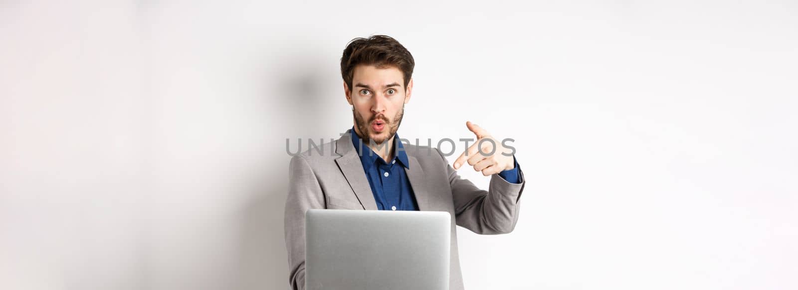 Excited handsome man asking to look here, pointing at laptop screen with amazed face, standing in business suit against white background.