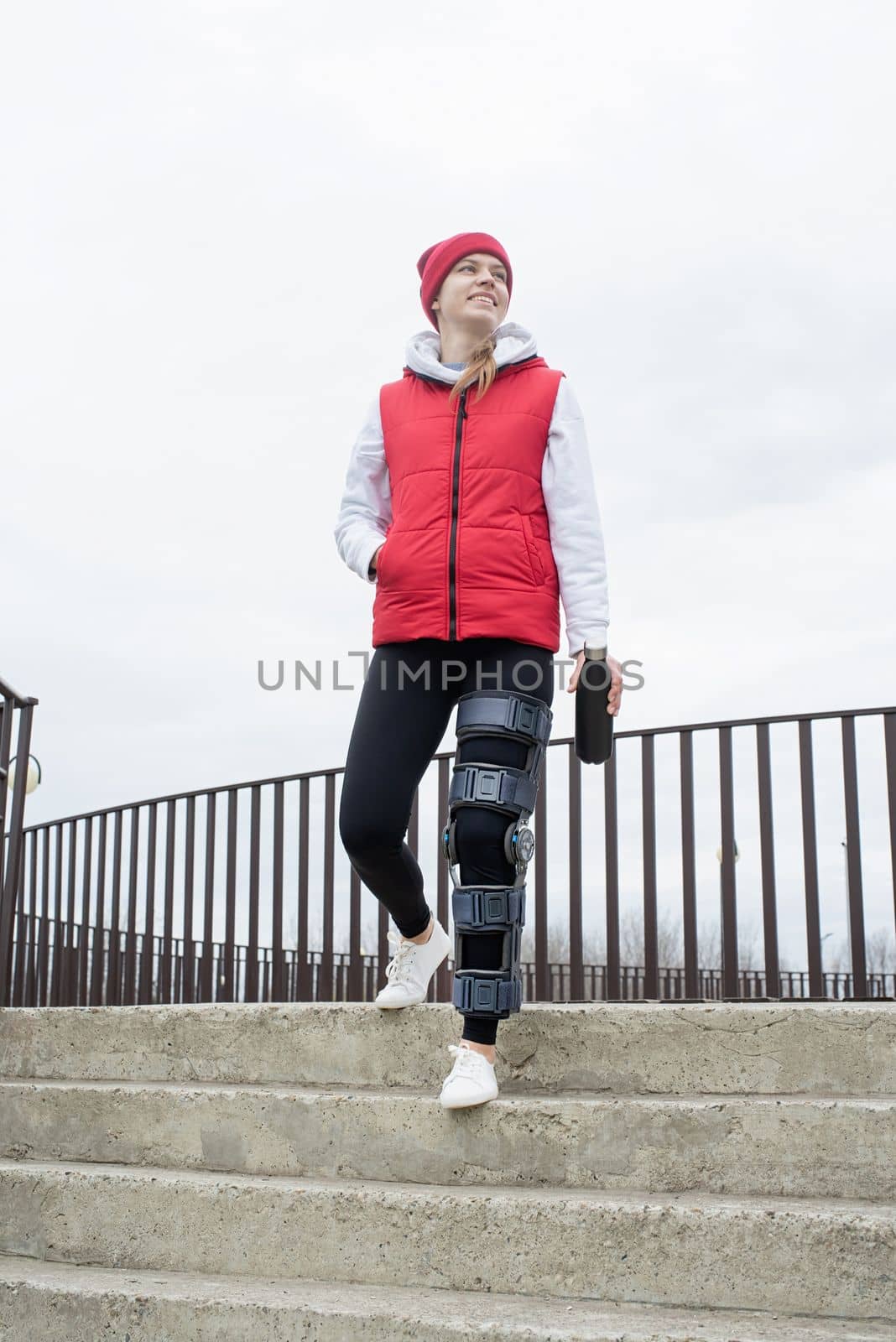 Woman wearing sport clothes and knee brace or orthosis after leg surgery, walking down the stairs in the park. Medical and healthcare concept.