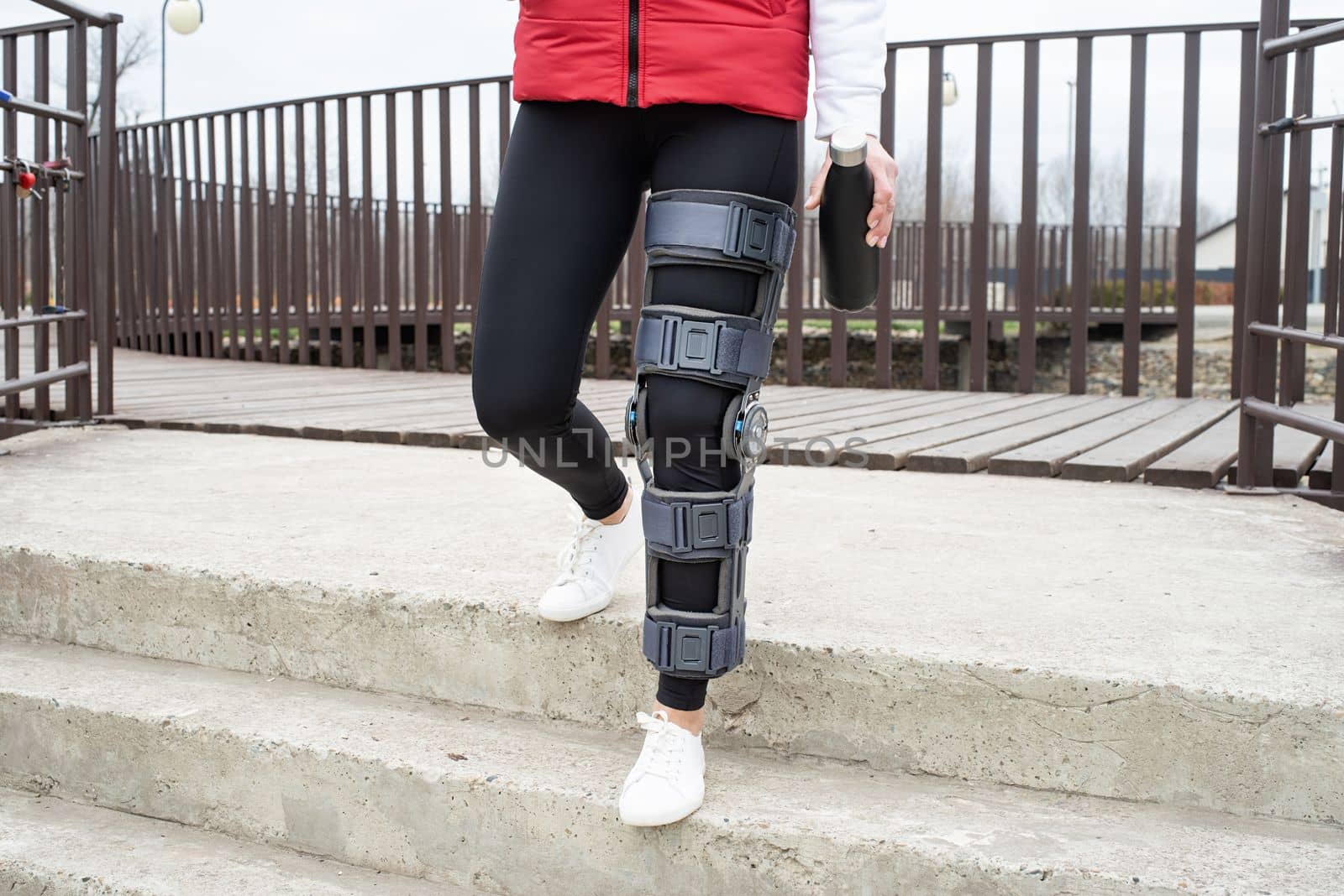 Woman wearing sport clothes and knee brace or orthosis after leg surgery, walking down the stairs in the park. Medical and healthcare concept.