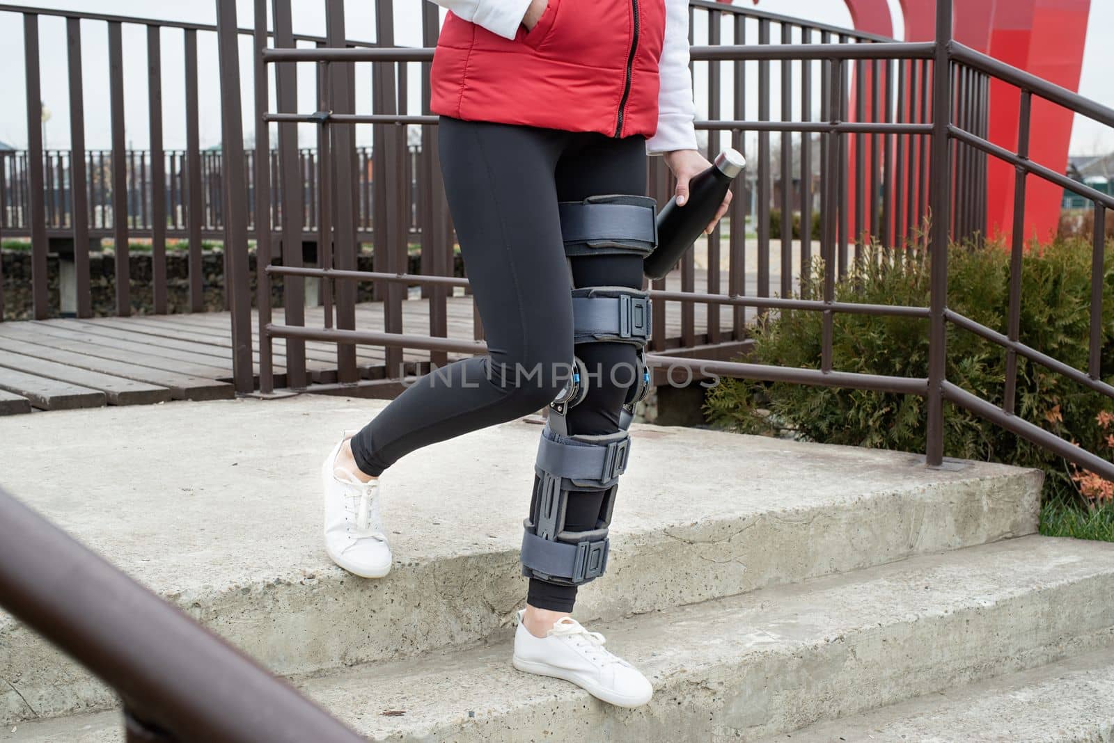 Woman wearing sport clothes and knee brace or orthosis after leg surgery, walking down the stairs in the park. Medical and healthcare concept.