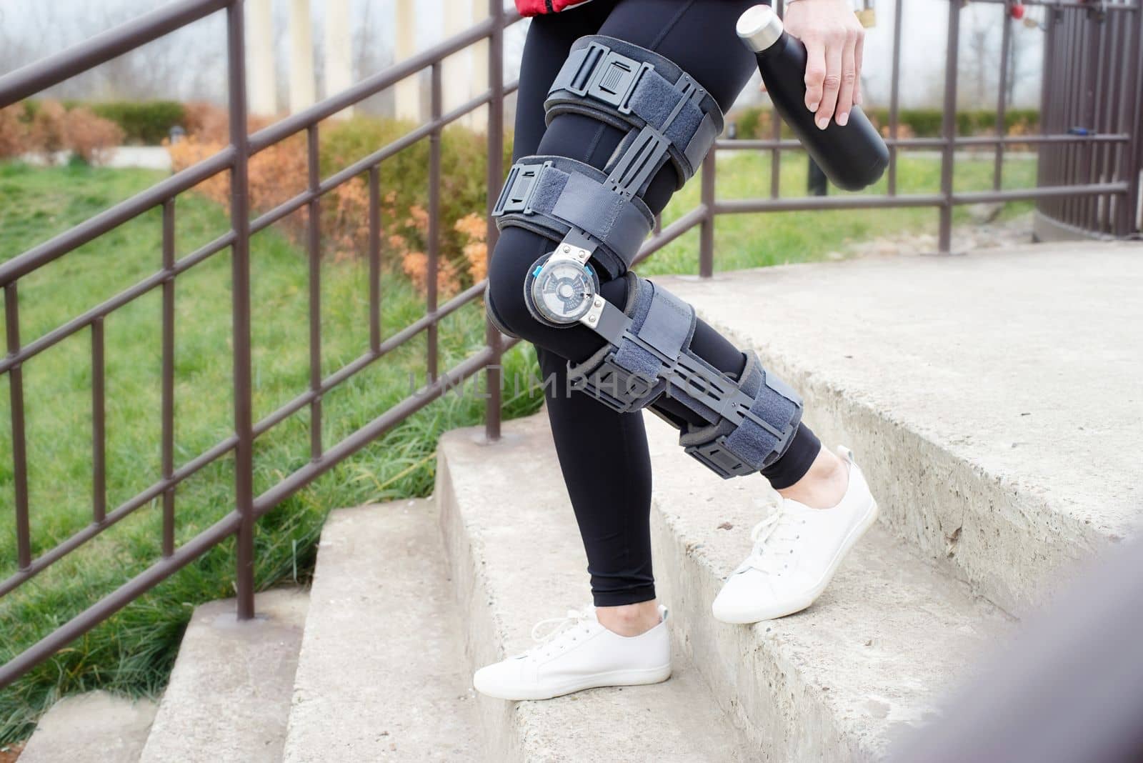 Woman wearing sport clothes and knee brace or orthosis after leg surgery, walking down the stairs in the park. Medical and healthcare concept.