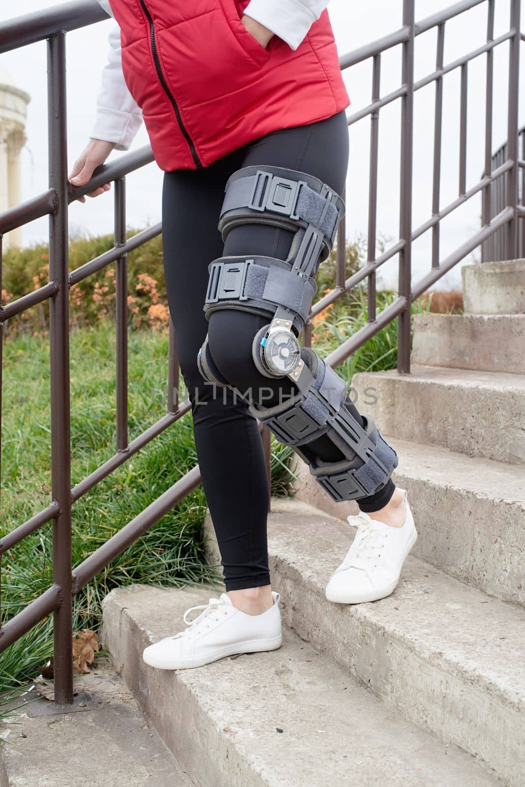 Woman wearing sport clothes and knee brace or orthosis after leg surgery, walking down the stairs in the park. Medical and healthcare concept.