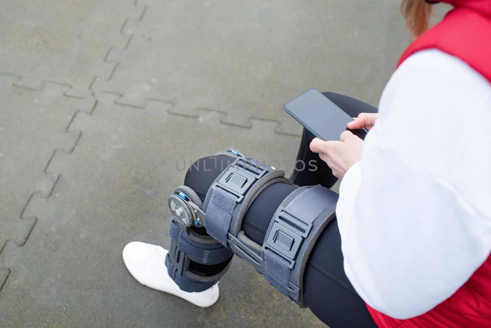 Woman wearing knee brace or orthosis after leg surgery working out in the park using smartphone . Medical and healthcare concept.