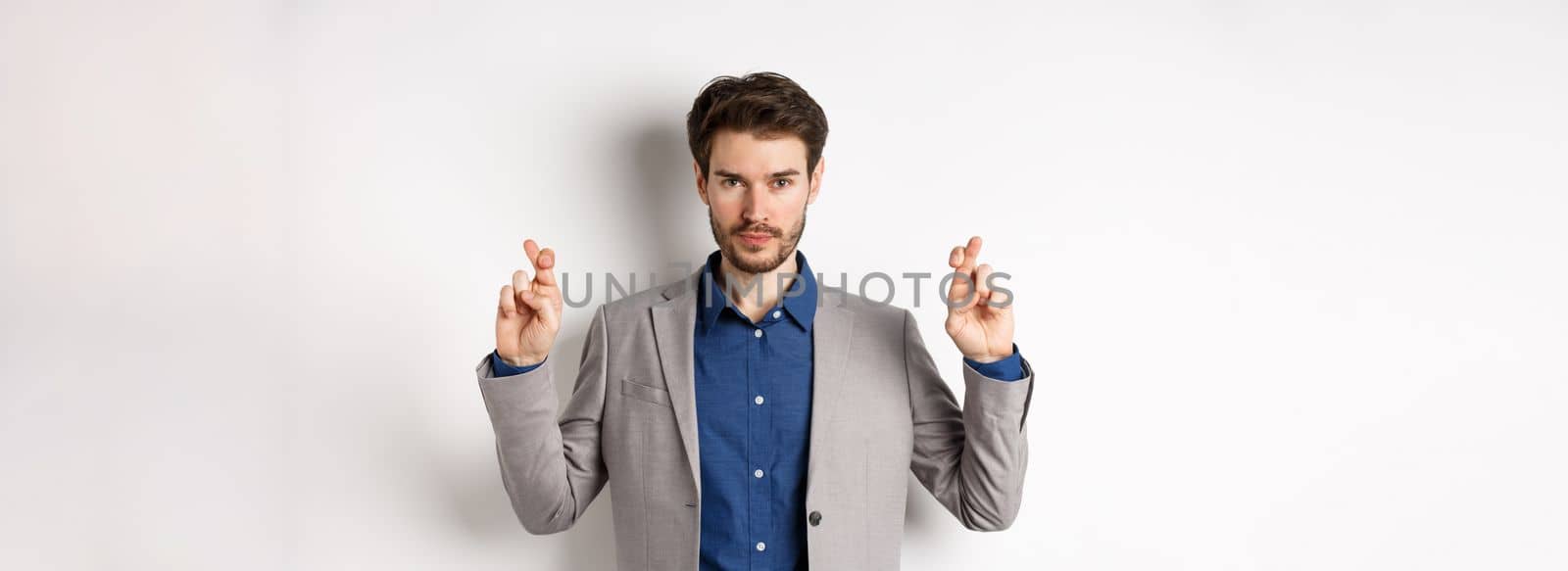 Hopeful man in suit cross fingers for good luck and looking confident in win, feeling determined, standing on white background.