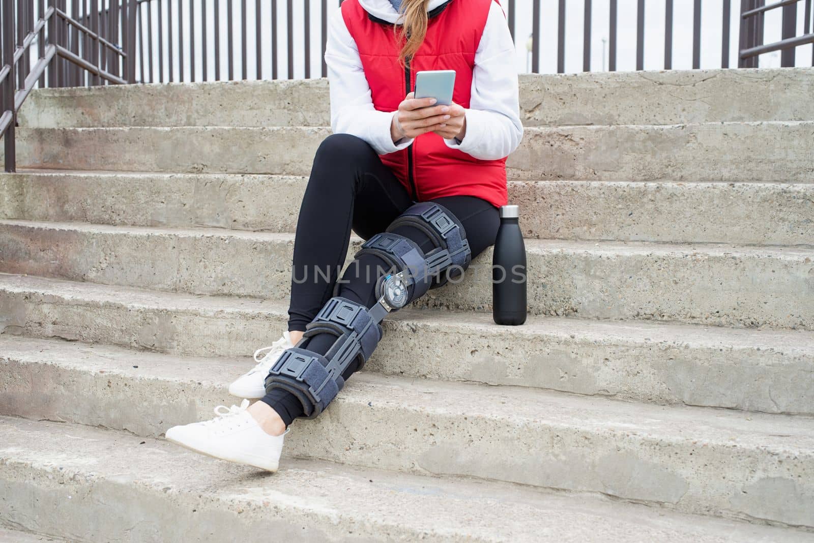 Woman wearing knee brace or orthosis after leg surgery, sitting on stairs ourdoors by Desperada