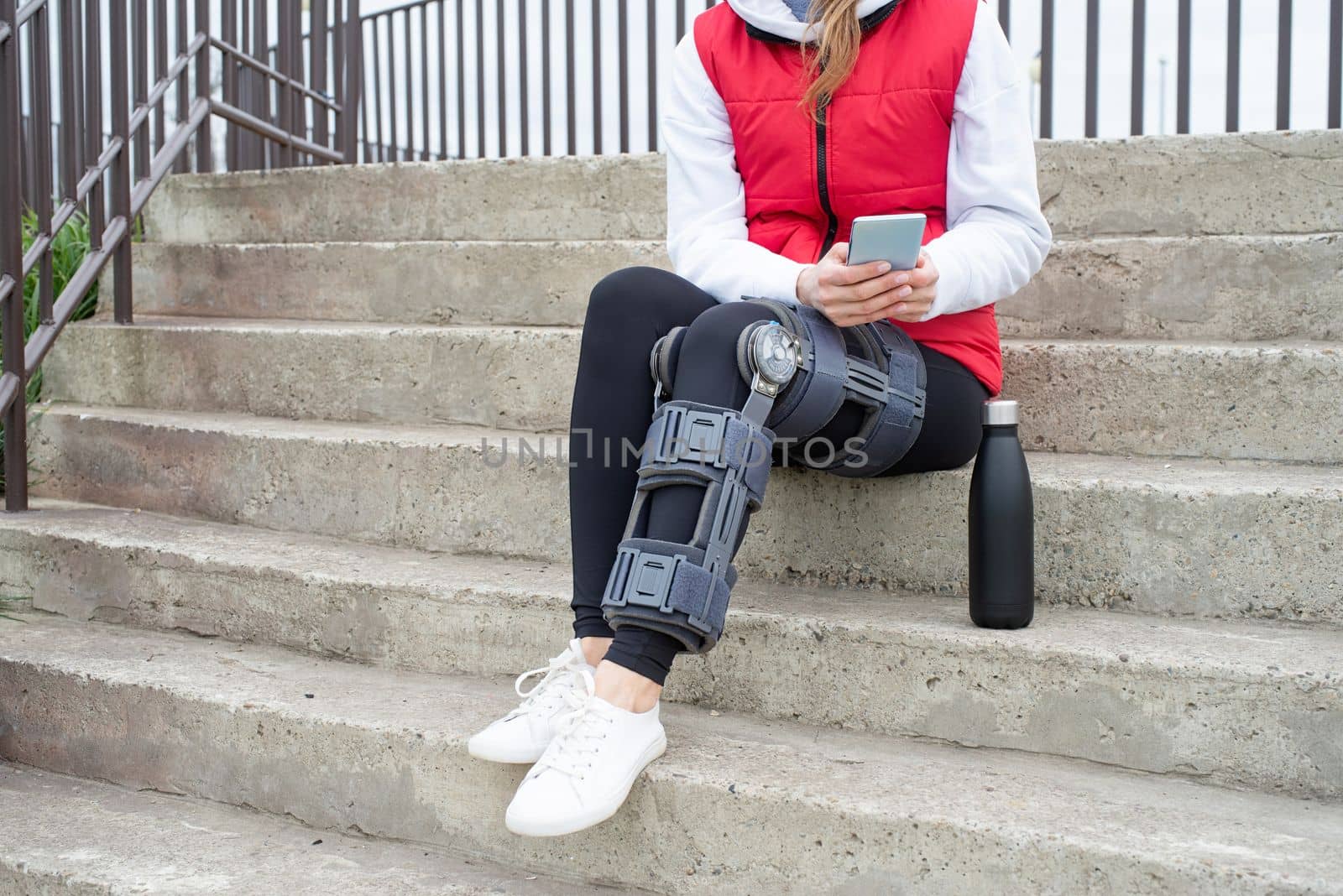 Woman wearing knee brace or orthosis after leg surgery, sitting on stairs ourdoors. Medical and healthcare concept.