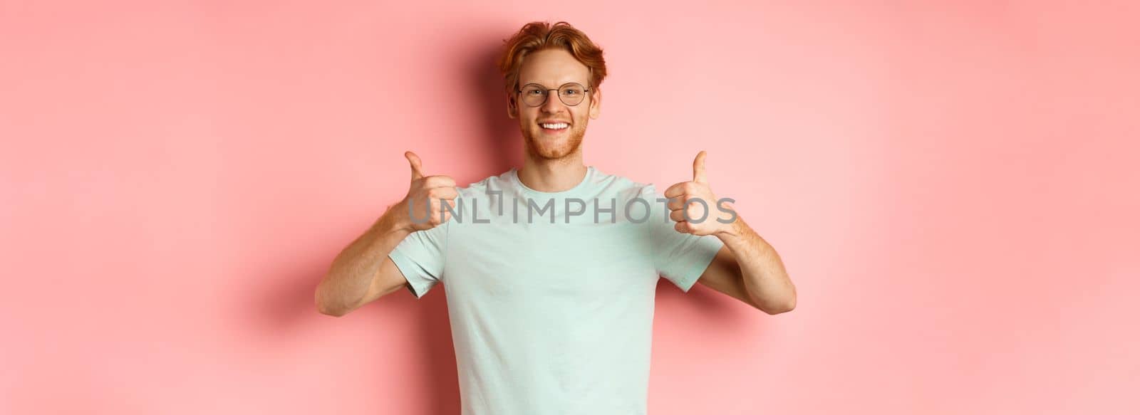Cheerful european man with red hair and beard, wearing glasses, showing thumbs-up and smiling in approval, praise something good, standing over pink background.