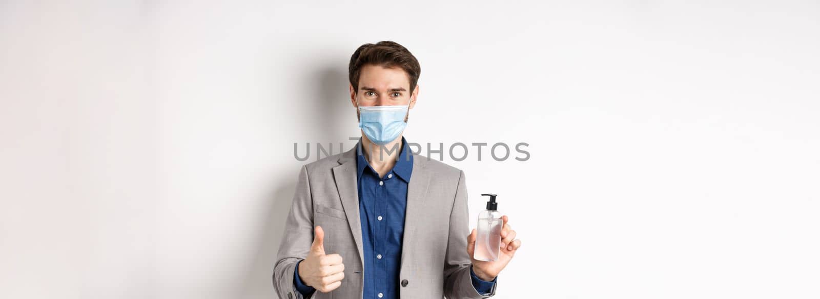 Covid-19, pandemic and business concept. Businessman in office suit and medical mask showing bottle of hand sanitizer and thumb up, recommend use antiseptic at work.