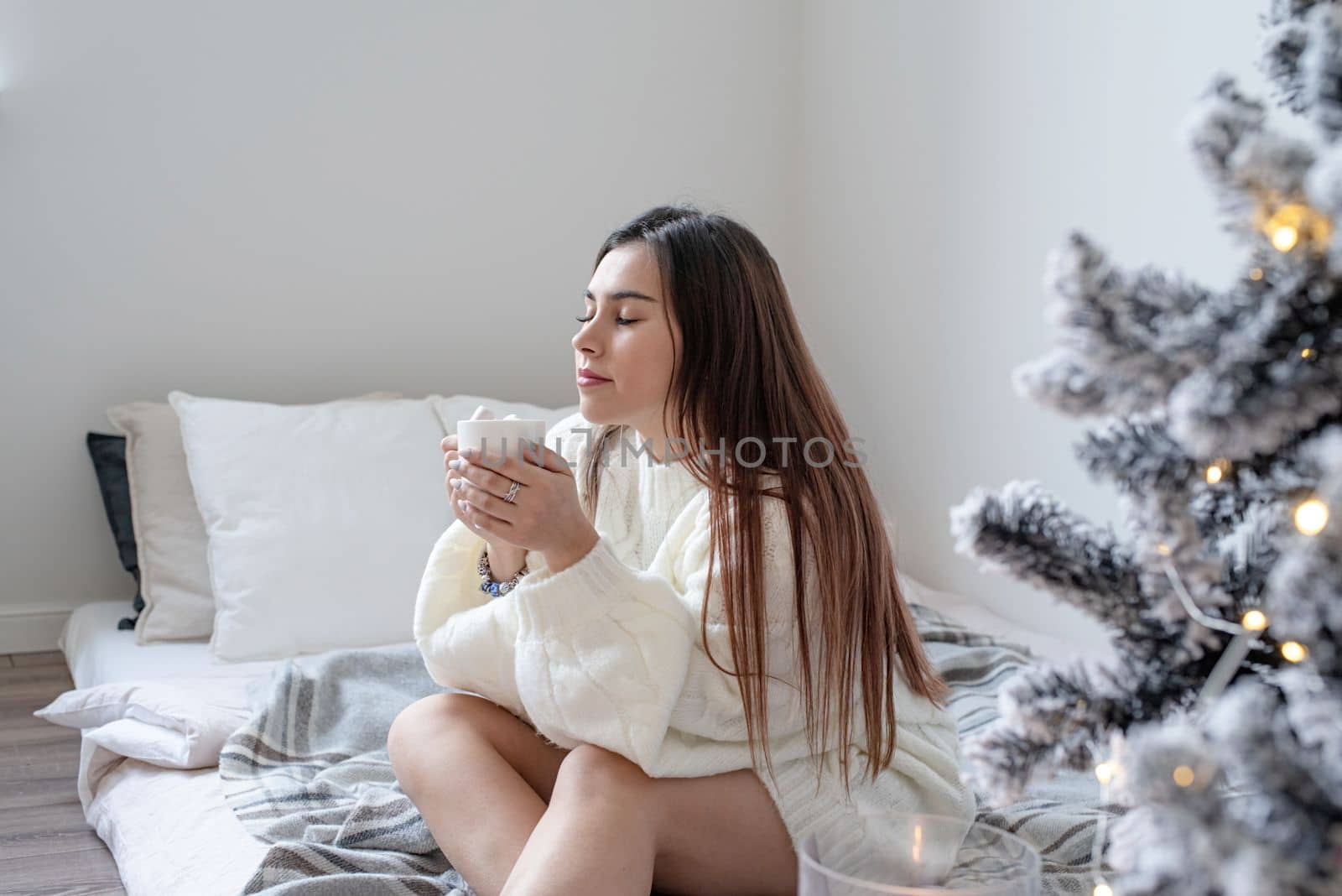 Merry Christmas and Happy New Year. Woman in warm white winter sweater lying in bed at home at christmas eve holding cup with marshmallows, fir tree behind